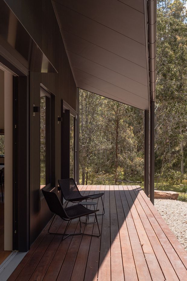 A narrow covered timber deck running alongside a dark-clad modern dwelling, with two black outdoor lounge chairs positioned against the wall. The deck has warm reddish-brown timber boards, a large sloping soffit overhead, slim structural posts, dark vertical wall cladding, and glazed openings connecting to the interior. A wooded natural setting is visible beyond the deck.