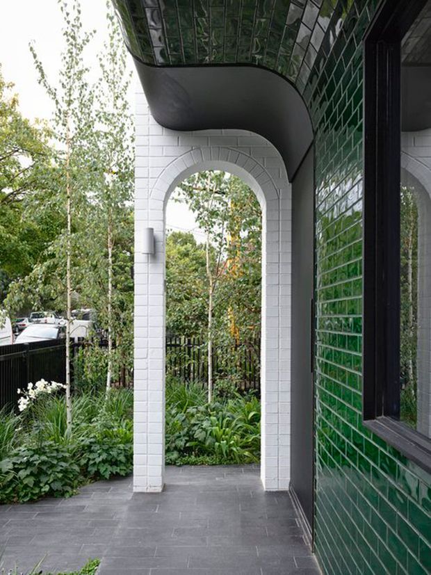 Covered exterior entry porch with a tall white tiled arched opening, dark grey tiled floor, and a striking glossy dark green tiled wall and curved soffit. Black-framed glazing reflects the entry area, while landscaped planting and a black fence are visible beyond the arch.