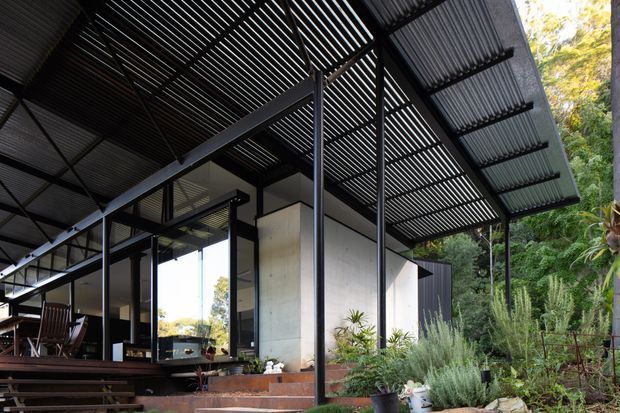 Exterior view of a contemporary pavilion-like house set within landscaped vegetation. The composition is dominated by a broad corrugated metal roof and a finely slatted pergola or screened roof structure supported by slender dark steel posts. Floor-to-ceiling glazing opens onto a timber deck with outdoor dining furniture, while pale rendered or concrete walls and dark vertical cladding form the building enclosure. Steps, raised garden beds, and dense planting frame the entrance and outdoor living area.