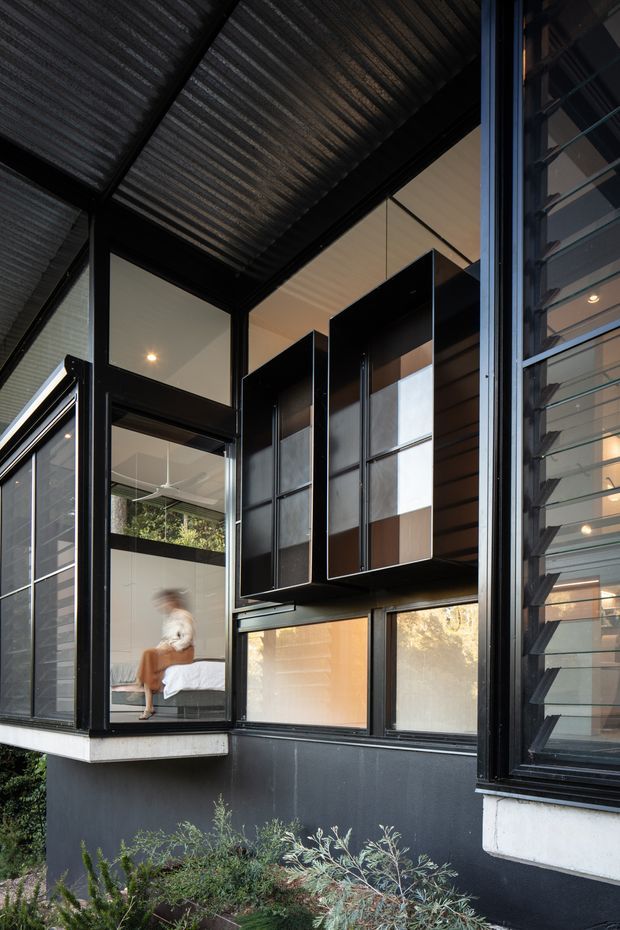 Exterior architectural photograph of a dark, modern multi-level facade with extensive black-framed glazing, projecting box windows, and a corrugated metal canopy or roof plane. Through the glazing, a bedroom with a seated person and a contemporary interior are visible. The facade includes louvred windows on the right, dark cladding, and a planted garden bed in the foreground.