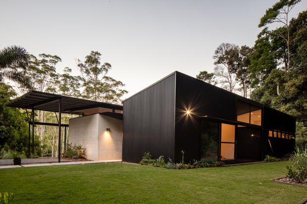 A low, modern black-clad house set within a lush landscaped garden. The angular composition features a large vertically ribbed dark facade, a broad flat-roofed carport or covered entry canopy, warm-lit floor-to-ceiling glazing, and a contrasting pale rendered wall. The building sits in a wooded, suburban or bush setting with a manicured lawn and planting beds, photographed at dusk.