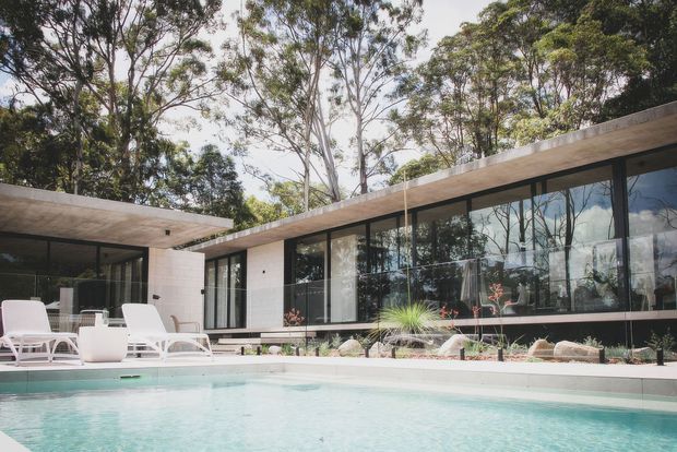 A low-slung modern residence set among mature trees, viewed from a swimming pool. The building has broad flat roof planes, extensive floor-to-ceiling glazing with dark frames, pale masonry walls, and a minimalist poolside terrace with white loungers and restrained landscaping.