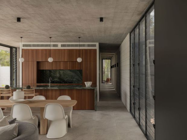 A refined open-plan dining and living space with a warm timber feature wall and integrated dark stone fireplace or media niche. A rounded timber dining table with sculptural cream chairs sits in the foreground, while full-height black-framed glazing lines the right side. The room has a concrete-look ceiling and large grey floor tiles, with a long hallway extending toward the rear. The overall design is restrained, contemporary, and material-focused.