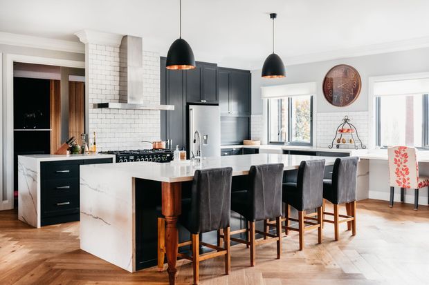 Bright, spacious open-plan kitchen and dining area with a large marble-look island, dark cabinetry, white subway-tile splashback, integrated stainless-steel appliances, and a long row of upholstered bar stools. Two black pendant lights hang above the island, while a dining nook and windows sit to the right. Warm herringbone timber flooring contrasts with the monochrome kitchen palette.