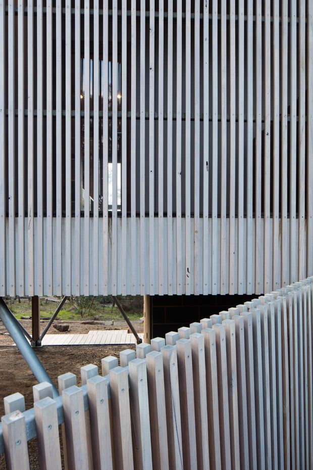 Close-up exterior architectural detail showing a raised building or structure clad in closely spaced vertical pale timber battens. The battens form a screened facade and continue into a curving or angled foreground fence, with glimpses of dark glazing and structural supports behind. The composition emphasizes repetition, texture, and contemporary lightweight construction.