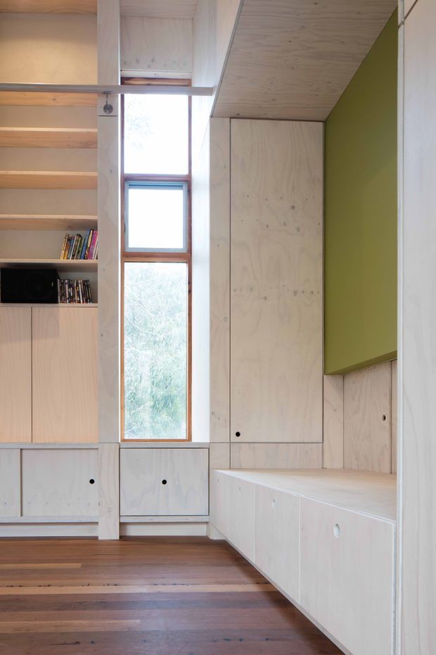 A compact, carefully integrated interior with pale plywood built-in cabinetry, shelving, drawers, a window seat, and a tall narrow timber-framed window. Open shelving holds books and a small television, while a muted olive-green wall panel provides contrast. The room has a warm timber floor and a calm, functional, space-efficient design.