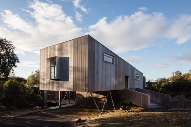 A compact modern elevated house set within a landscaped site, clad in vertically ribbed timber boards. The simple elongated box has a sharply pitched or sloping roofline, narrow horizontal windows, and a projecting window with a dark louvered or shutter-like panel. The building is raised on slender angled supports, with a timber access ramp and deck leading to the entrance. Warm low-angle sunlight emphasizes the textured facade and structural supports.