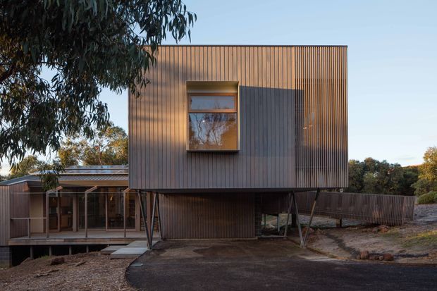 Exterior view of a contemporary elevated house with a box-like upper storey clad in vertical timber battens. The building is supported on slender angled posts above a lower glazed level and deck, with a dark paved driveway in front and mature trees surrounding the site.