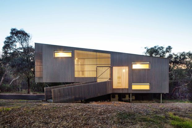 A compact contemporary elevated dwelling set among native trees, photographed at dusk. The rectangular building has dark vertically ribbed cladding, a broad warm-lit glazed central section, narrow horizontal windows, and a timber access ramp leading to a simple entry door. The structure appears raised on short supports and sits in a natural bush setting.