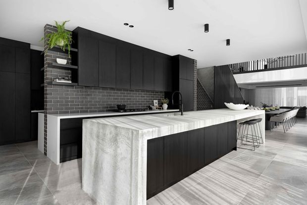A dramatic open-plan kitchen with a long waterfall island, dark vertically grained cabinetry, charcoal brick-look splashback, and a pale veined stone benchtop. The kitchen opens toward a dining and living area, with a black metal stair and upper-level balustrade visible in the background. Minimal black cylindrical and recessed lighting complements the monochromatic scheme.