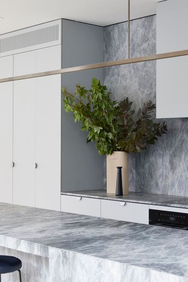 A contemporary kitchen with extensive white built-in cabinetry, a grey marble-look island and benchtop, and a matching textured stone-look splashback. A black appliance is integrated beneath the rear cabinetry, while a large vase with leafy branches adds natural contrast. A slim timber-toned horizontal feature element spans the upper portion of the room.