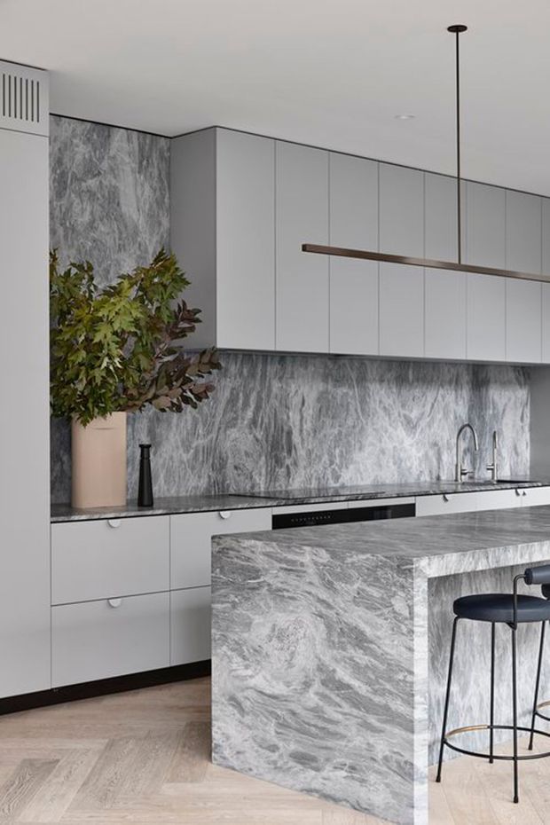 Contemporary kitchen featuring pale grey flat-panel cabinetry, a dramatic grey-and-white marble-look stone backsplash and waterfall island, integrated appliances, black plumbing fixtures, and a slim linear timber pendant. Light herringbone flooring and restrained styling create a refined, monochromatic space.