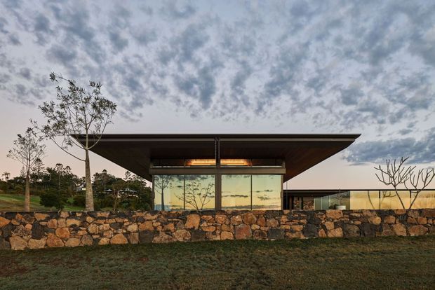 Single-storey contemporary pavilion-style house viewed from the exterior at dusk, with a broad low-pitched projecting roof, extensive floor-to-ceiling glazing, dark structural framing, and a long low adjoining wing. A dry-stacked natural stone boundary wall and landscaped lawn sit in the foreground, with trees and a partly cloudy sky beyond.