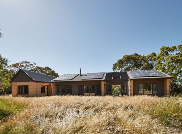 Single-storey contemporary residential house set in a rural landscape, with low-pitched gabled roof forms, extensive rooftop solar panels, timber-toned cladding, dark window and door frames, and broad glazed openings. The home is surrounded by tall dry grasses and mature trees under a clear blue sky.