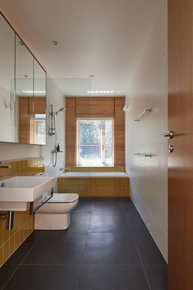 A compact contemporary bathroom viewed from the doorway, featuring a freestanding bathtub beneath a window, wall-mounted shower fittings, a white wall-hung basin, toilet, large frameless mirror, white wall tiles, mustard ochre tiled bathtub surround, dark charcoal floor tiles, and warm timber window and door finishes.