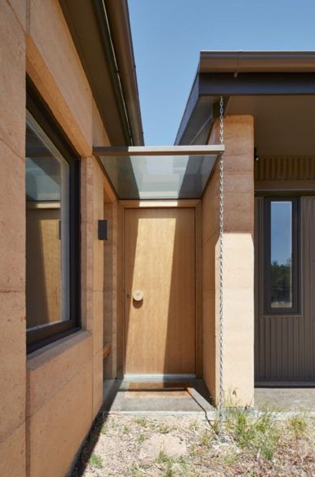 A close exterior view of a contemporary residential entry. A timber-clad front door sits beneath a translucent glass canopy supported by a slender metal frame and rain chain. Warm earthen-toned masonry or rendered wall surfaces contrast with dark window frames and corrugated dark cladding on the adjoining structure. The entry is reached by a small concrete threshold and sits beside sparse landscaping.