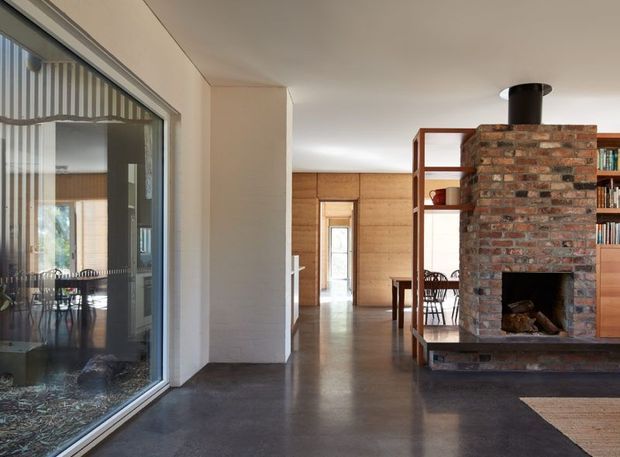 A warm, open-plan residential interior with polished dark concrete flooring, timber-lined walls, and a substantial exposed brick fireplace with a black flue. A timber-framed open shelving partition separates the fireplace and dining area from the circulation space. Full-height glazing on the left looks into a dining or sunroom, while a timber-lined room and doorway extend into the background.