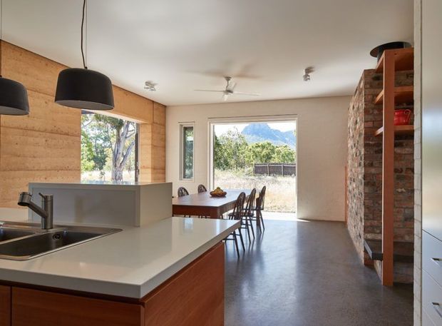 Open-plan kitchen and dining area with a white island benchtop, timber cabinetry, timber wall lining, and an exposed brick shelving feature. A dining table sits beside large glazing and a sliding glass door opening to an outdoor view, with a mountain visible beyond. The room has polished concrete flooring, simple contemporary lighting, and a ceiling fan.