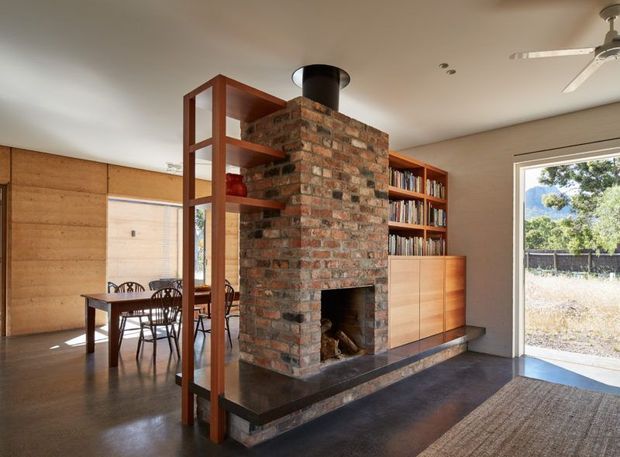Warm open-plan living and dining space centred on a freestanding exposed-brick fireplace with a black flue and hearth. Built-in timber cabinetry and shelving, a timber-framed open display shelf, dining table, and extensive book storage define the room. A large glazed opening provides views outdoors, while a ceiling fan and neutral plaster ceiling complete the understated interior.
