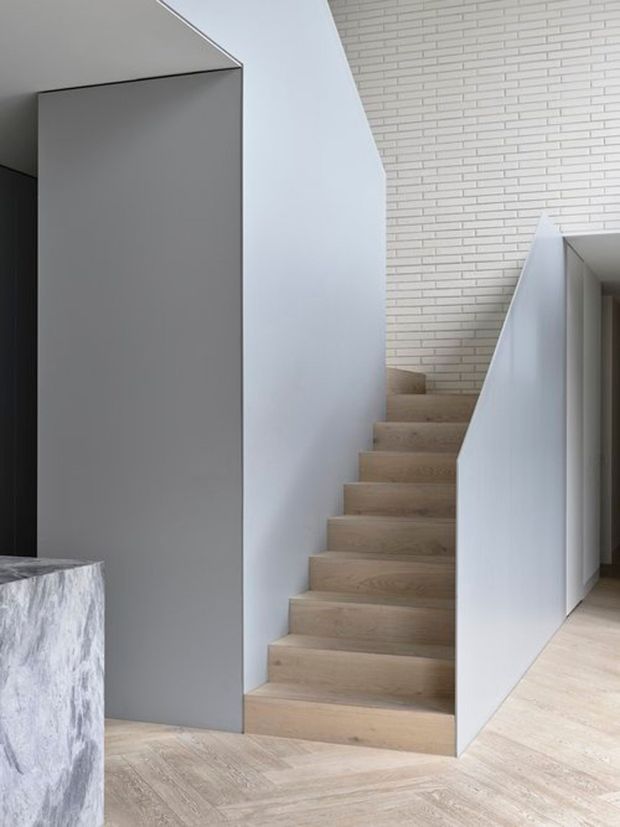 A minimalist interior stairway with pale oak-toned timber stairs and flooring, crisp white walls, a smooth grey partition, and a white painted brick feature wall. The stair has a clean, enclosed geometric form leading toward a hallway, with a light neutral palette and restrained contemporary detailing.