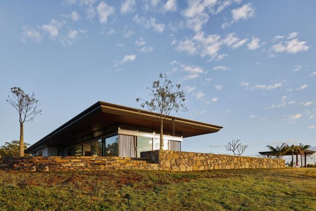 Low-slung contemporary single-storey building set in a landscaped, gently sloping site. The structure features a broad projecting flat roof, extensive floor-to-ceiling glazing, dark slender framing, warm timber soffits, and a low irregular natural-stone wall running across the foreground. Sparse landscaping and distant palms surround the building under a clear blue sky.
