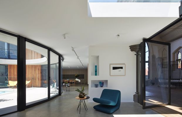 A bright contemporary interior lounge and circulation space with polished concrete flooring, white walls and ceiling, extensive black-framed glazing, and a large roof skylight. A sculptural blue lounge chair and small side table sit in the foreground, while a dining or kitchen area is visible beyond. Open glazed doors connect the room to an exterior courtyard and a darker covered veranda with ornate existing architectural elements.