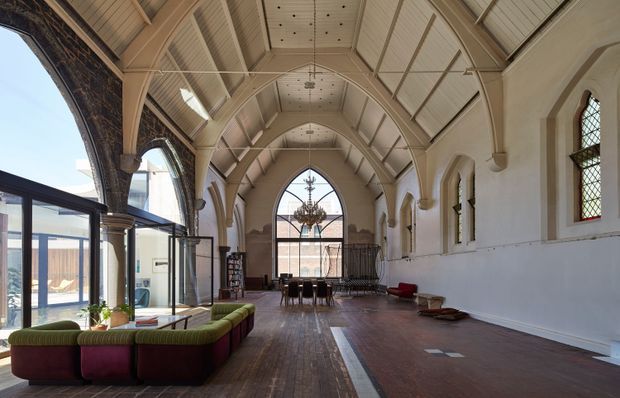A spacious adaptive interior within a historic church-like hall, featuring a tall vaulted and ribbed ceiling, pointed arched windows, exposed dark stone arch surrounds, timber flooring, and a large central arched glazed opening. Contemporary lounge seating and a dining or meeting arrangement occupy the open nave-like space, while extensive modern glazing connects to an adjoining bright area on the left. The atmosphere is dramatic, refined, and airy, combining heritage architectural details with contemporary furnishings.