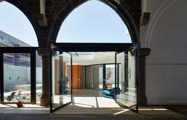 View through a historic stone arched colonnade into a bright contemporary interior. Large black-framed glazed folding or pivot doors stand open, creating a strong transition between the masonry arcade and the modern room beyond. The interior features pale flooring, dark-framed glazing, warm timber wall panelling, and a sculptural blue lounge chair, with a small courtyard or planted area visible to the left.