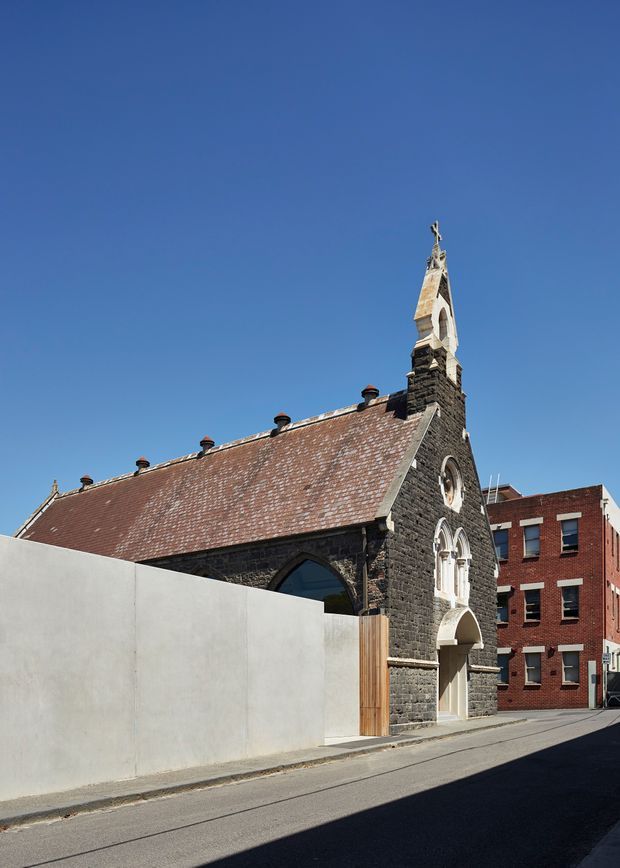 Street-level exterior view of a historic Gothic Revival stone church or chapel in an urban setting. The building has dark masonry walls, a steep weathered reddish tiled roof with multiple small chimneys, pointed-arch windows and entry, ornate pale stone tracery, and a narrow bell tower topped with a cross. A contemporary pale boundary wall and timber gate sit in front, with a red-brick multi-storey building adjacent.