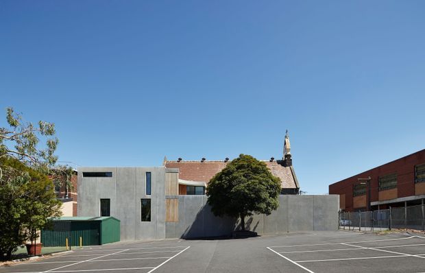 Wide exterior view of a contemporary grey concrete building beside a paved marked parking area. The facade has simple rectangular openings, a vertical timber-clad section, and a low green service structure, with a brick building and church-like roof element visible behind. Clear blue sky and mature trees surround the site.