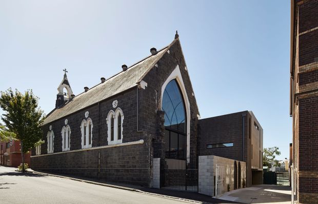 Exterior view of a historic Gothic Revival church constructed in dark masonry, with steep gabled roof, pointed-arch windows, a small bell tower, and a large contemporary glazed entrance addition. The church sits within a dense urban streetscape alongside modern brick buildings.