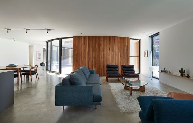 A bright, open-plan modern living and dining area with polished concrete flooring, white walls, extensive black-framed glazing, and a prominent curved vertical timber feature wall. The living zone contains a deep blue sofa, matching armchair, two dark lounge chairs, a glass coffee table, and a neutral rug. A dining table and chairs sit beside a minimalist kitchen island, while a long recessed wall shelf displays small decorative objects.