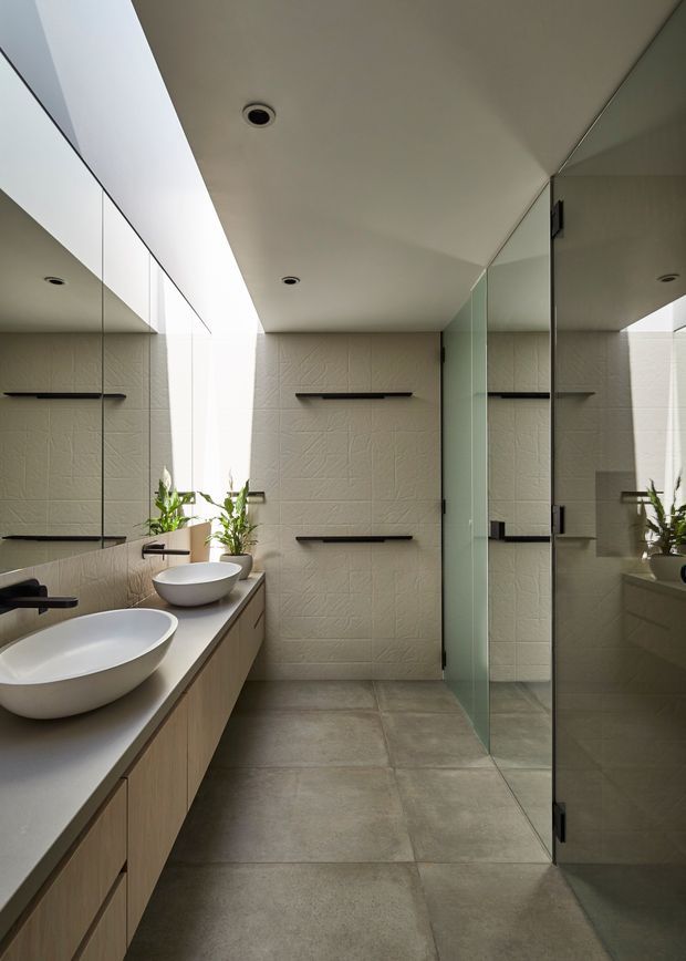 A contemporary bathroom featuring a long floating timber-toned vanity with two white vessel basins, dark wall-mounted tapware and towel rails, large grey floor tiles, textured pale wall tiles, and a clear glass shower enclosure. Full-height mirrors expand the narrow room, while a skylight or clerestory opening brings in natural light. Small plants add a natural accent.
