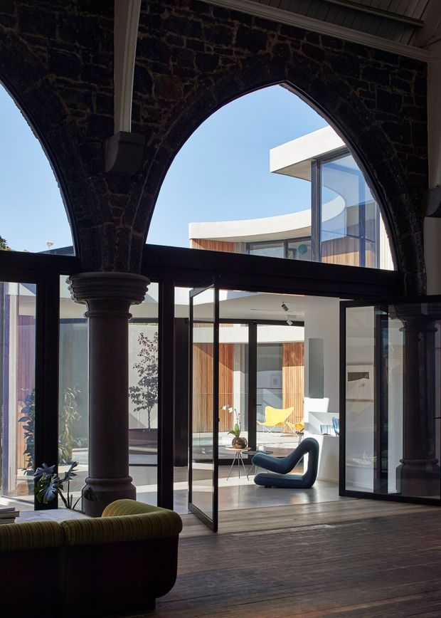 A dramatic interior view through a heritage-style dark masonry arcade with pointed arches, substantial columns, and dark timber flooring. Black-framed glazed doors open toward a bright contemporary courtyard living space featuring timber wall cladding, white walls, sculptural modern seating, and a curved flat-roof form beyond. The image contrasts historic architectural fabric with a light-filled contemporary addition.