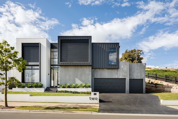Street-facing photograph of a contemporary multi-level residence with a strong geometric facade. The home combines smooth light-grey rendered concrete-like walls, dark vertical metal cladding, large dark-framed windows with external louvre or roller-shutter screens, and two dark garage doors. A broad paved driveway, low landscaped frontage, retaining wall, fencing, and a visible house number complete the suburban exterior.