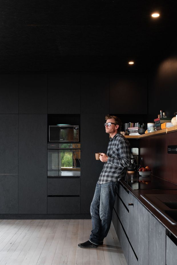 A dramatic, dark-toned contemporary kitchen with full-height matte black cabinetry, integrated wall ovens, a long dark benchtop, open shelving, and warm recessed ceiling lights. A person stands at the island or workbench holding a mug. Pale timber flooring provides contrast against the predominantly black and charcoal fit-out.