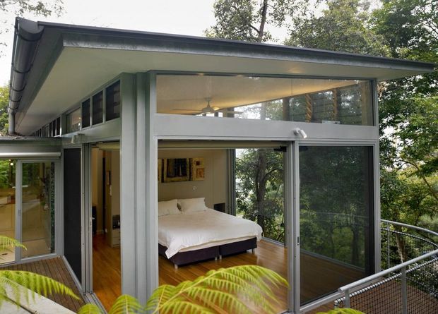Exterior view of a raised, tree-surrounded modern home with a partially enclosed bedroom opening onto a narrow deck or balcony. The structure has broad low-pitched roof eaves, extensive floor-to-ceiling glazing, grey metal-framed windows and doors, and timber flooring visible inside. A bed is clearly visible through the open glass wall, with dense foliage surrounding the building.