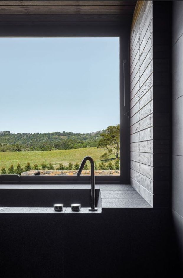 A dramatic, minimalist bathroom interior focused on a deep dark freestanding bathtub beneath a large picture window. A tall black gooseneck tap and two controls sit on the stone bath surround. Dark timber ceiling boards, dark wall finishes, and a contrasting textured light-grey masonry wall frame a rural green landscape outside.