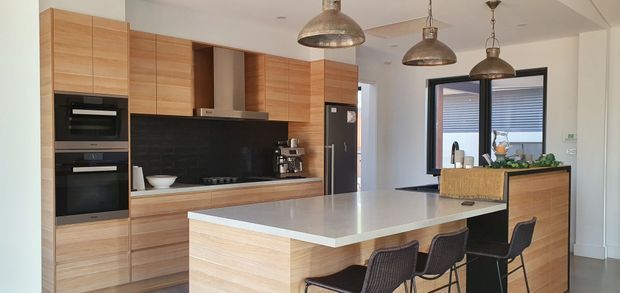 A contemporary open-plan kitchen with extensive light timber cabinetry, integrated black appliances, a large pale stone island with bar seating, black-framed windows, and three industrial-style pendant lights. A raised timber-clad counter or servery with plants is visible beyond the island.