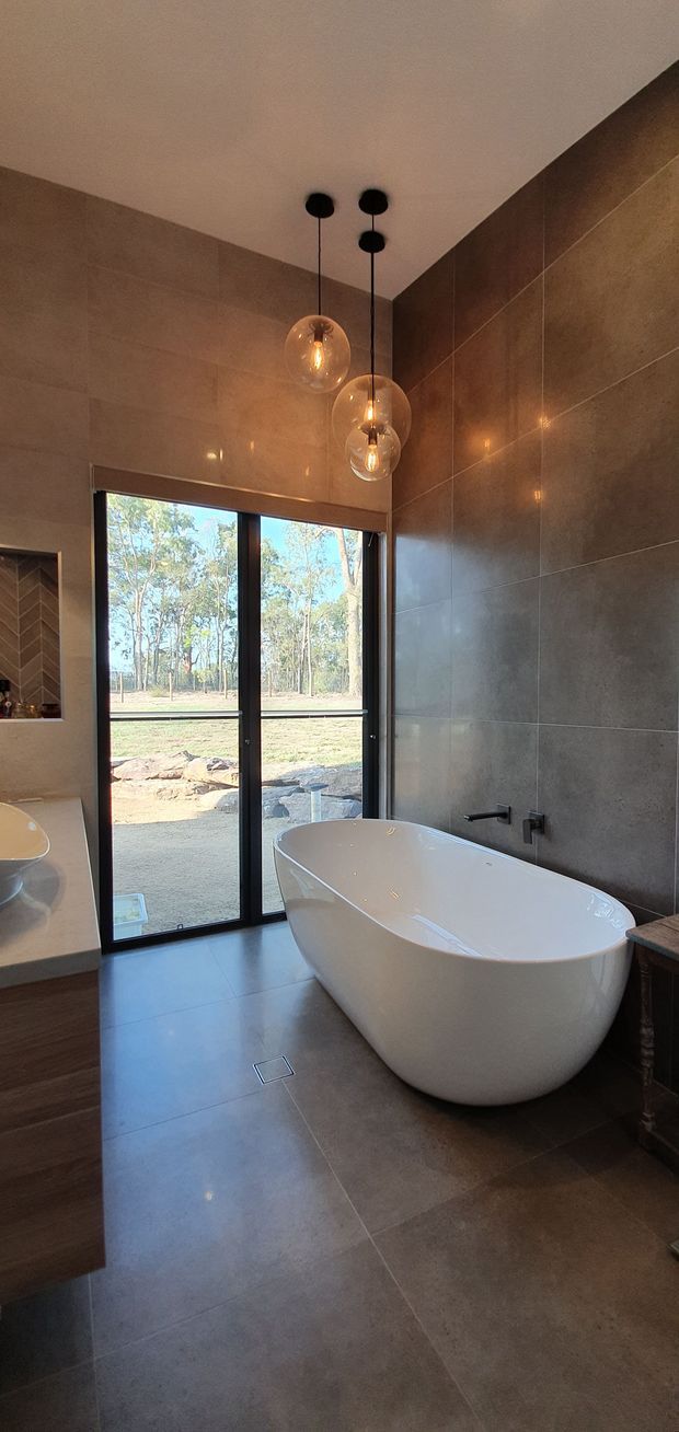 A contemporary bathroom with large-format warm grey-brown stone-look tiles covering the walls and floor. A freestanding white oval bathtub sits beneath three suspended clear glass pendant lights with warm bulbs. A dark-framed glazed door opens to a wooded outdoor setting, while timber-toned vanity cabinetry and a recessed herringbone-tile niche are visible at the left.