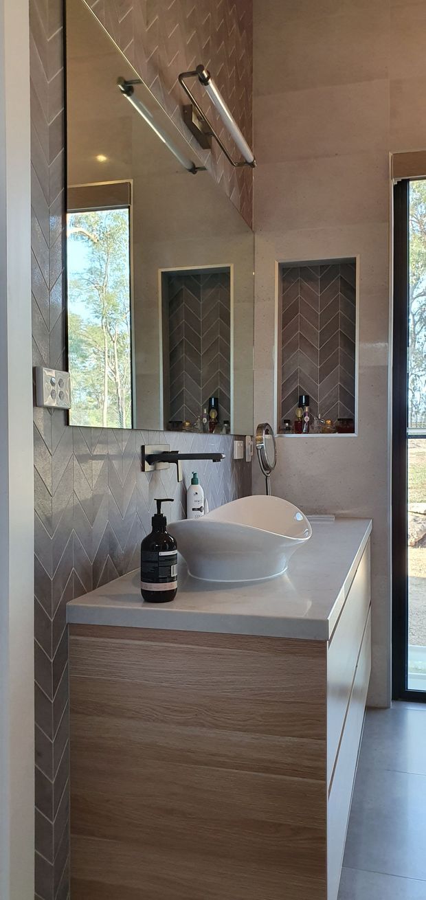 A contemporary bathroom vanity area with a freestanding white vessel basin on a pale stone benchtop, timber-look cabinetry, wall-mounted dark metal tapware, a large mirror, and patterned chevron wall tiles. A tall window provides natural light and views of trees.