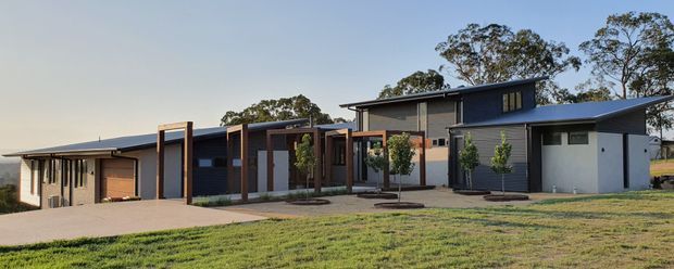 A wide exterior view of a low-profile contemporary single-storey residence. The home combines dark grey rendered walls, extensive timber-framed glazing and screens, a broad projecting roofline, and a partly raised central roof volume. A paved driveway and landscaped lawn sit in front of the house, with mature trees in the background.