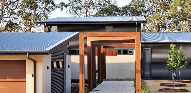 Contemporary single-storey residential exterior with dark horizontal cladding, low-pitched metal roofing, rendered light walls, and a prominent timber-framed covered walkway or entry pergola. The composition includes multiple connected roof forms, a garage-like volume at left, and landscaped planting around the entry.