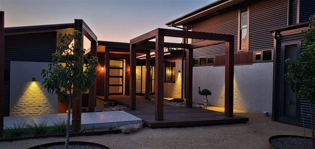 A contemporary residential courtyard and entry facade photographed at dusk. The composition features dark horizontal cladding, warm timber-framed portals and a covered timber entry walkway, rendered masonry walls with integrated lighting, textured paving, gravel landscaping, and concealed warm illumination.