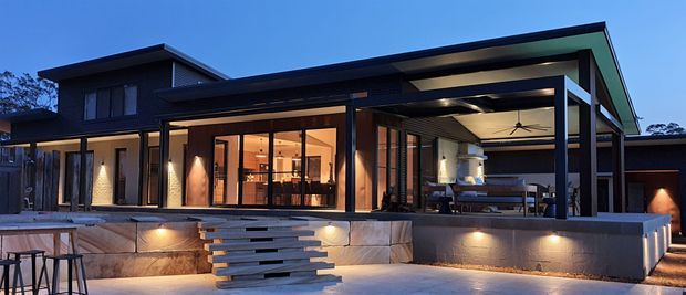 Contemporary two-storey residential home photographed at dusk, with a dark-clad upper level and expansive glazed living areas opening onto a covered outdoor entertaining terrace. The terrace features a pitched roof extension, ceiling fan, built-in fireplace or cooking area, and outdoor dining/lounge furniture. Broad concrete retaining walls, floating timber-look steps, and warm architectural lighting define the foreground.