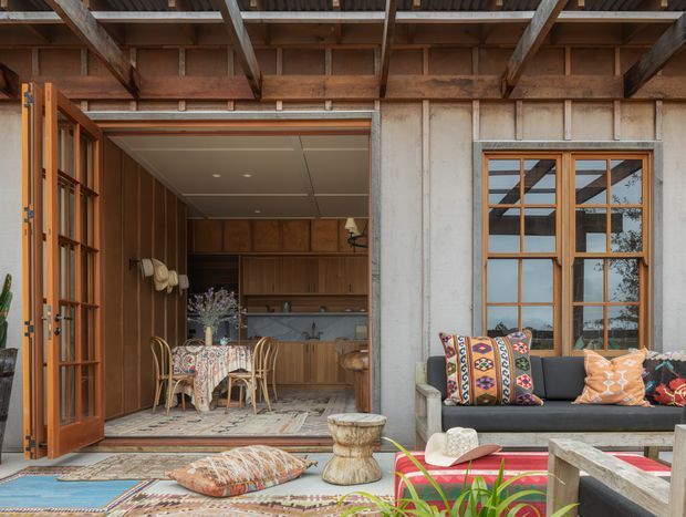 A rustic covered outdoor entertaining area opening into a warm timber-lined kitchen and dining space. Exposed timber roof beams, timber-framed glazed doors and windows, concrete wall panels, patterned rugs, a dark sofa with colourful cushions, and a small outdoor coffee table create a relaxed indoor-outdoor setting.