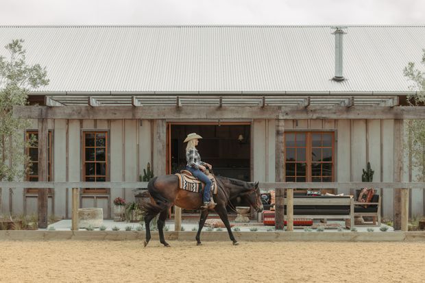 Front elevation of a rustic, barn-like single-storey building with weathered vertical timber cladding, exposed timber framing, a large corrugated metal pitched roof and chimney. Multi-pane timber-framed windows and a central open doorway reveal a dark interior with rustic furnishings. A covered veranda with timber posts and railing runs along the facade, while a rider on horseback crosses the sandy foreground.