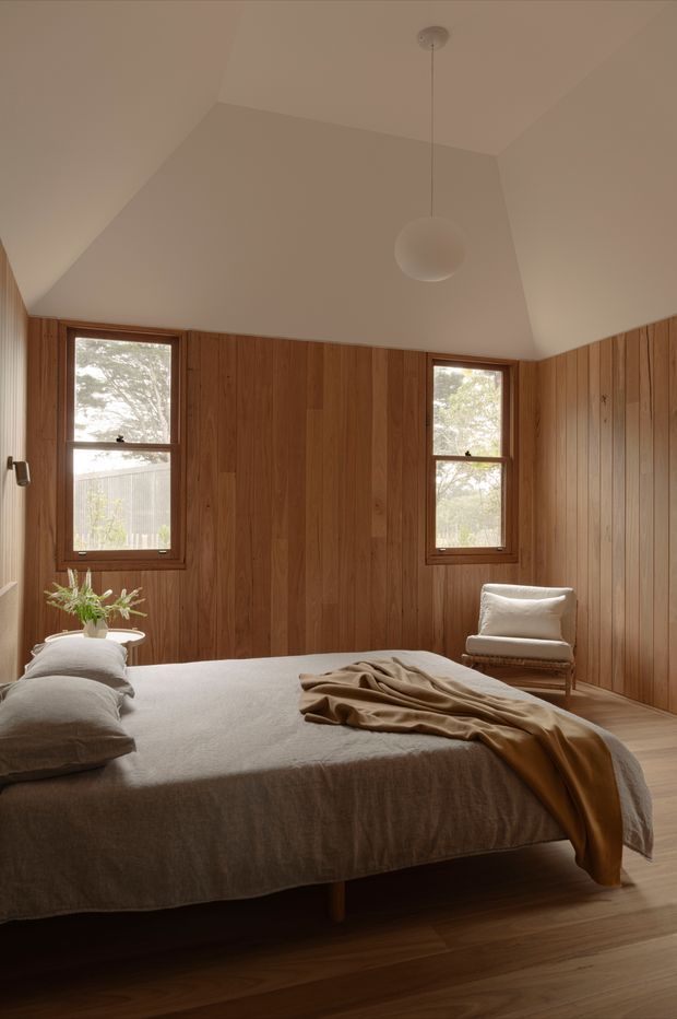 A warm, serene bedroom with full-height vertical timber wall panelling, timber flooring, two simple framed windows, and a sculptural white pendant light beneath an angled ceiling. A low-profile bed with neutral linen bedding occupies the foreground, with a small upholstered armchair and a vase of greenery adding restrained furnishings. The room has a natural, minimalist character with abundant warm wood and soft beige textiles.