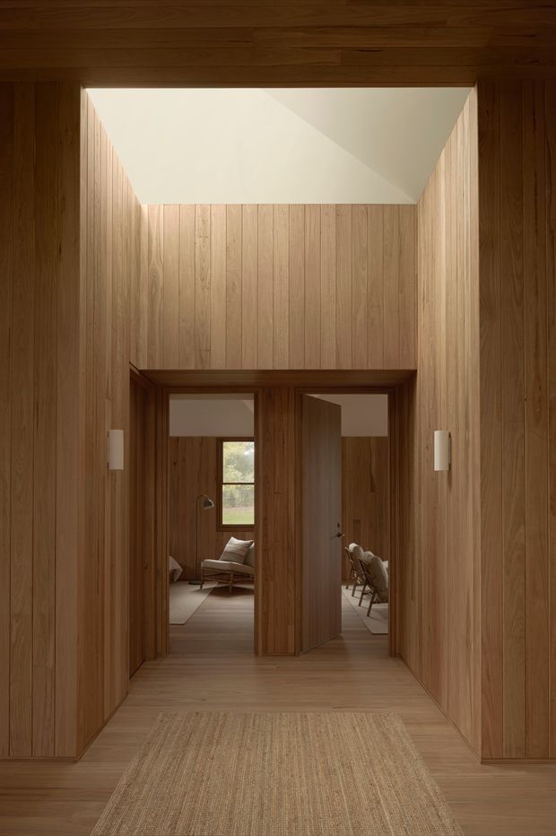 A warm, timber-lined residential hallway or entry corridor with full-height vertical wood panelling, matching timber floor and ceiling, and a large rectangular skylight or clerestory opening that brings in soft daylight. At the end of the corridor, open timber doors reveal adjoining furnished rooms, including seating and a dining area. A neutral woven runner leads through the space, while slim white wall lights provide understated illumination.