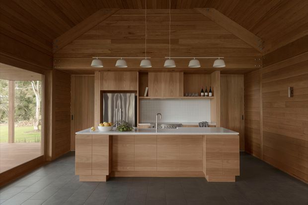 A warm timber-lined kitchen with a pitched, exposed timber ceiling and integrated timber cabinetry. A large central island with a pale stone benchtop faces the camera, while the rear wall contains a sink, tiled splashback, shelving, and concealed appliances. A row of white pendant lights hangs above the island, and full-height glazing on the left opens views to the surrounding landscape.